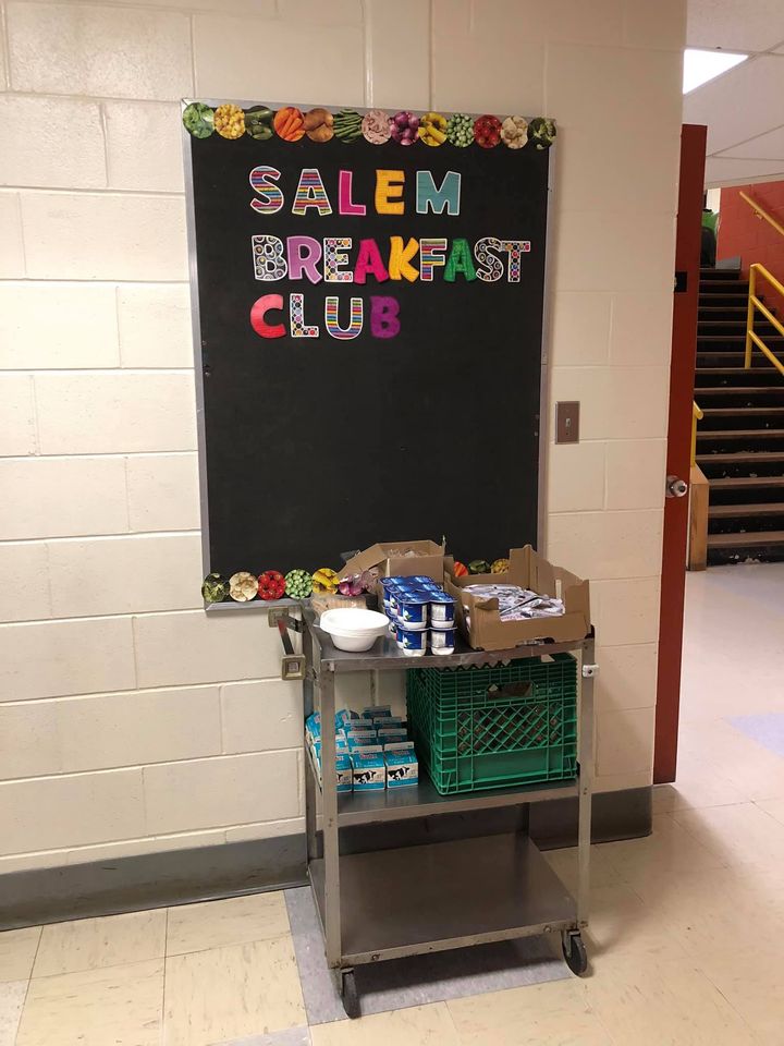 A black bulletin board is on a white brick wall indoors. "SALEM BREAKFAST CLUB" in multicoloured letters is posted to the board. There is a boarder on the top and bottom of the board that is a garland made of circular images of food. A grey cart with wheels is in front of the board. On the cart is plastic bowls, yogurt, disposable cutlery, and small cartons of milk. 