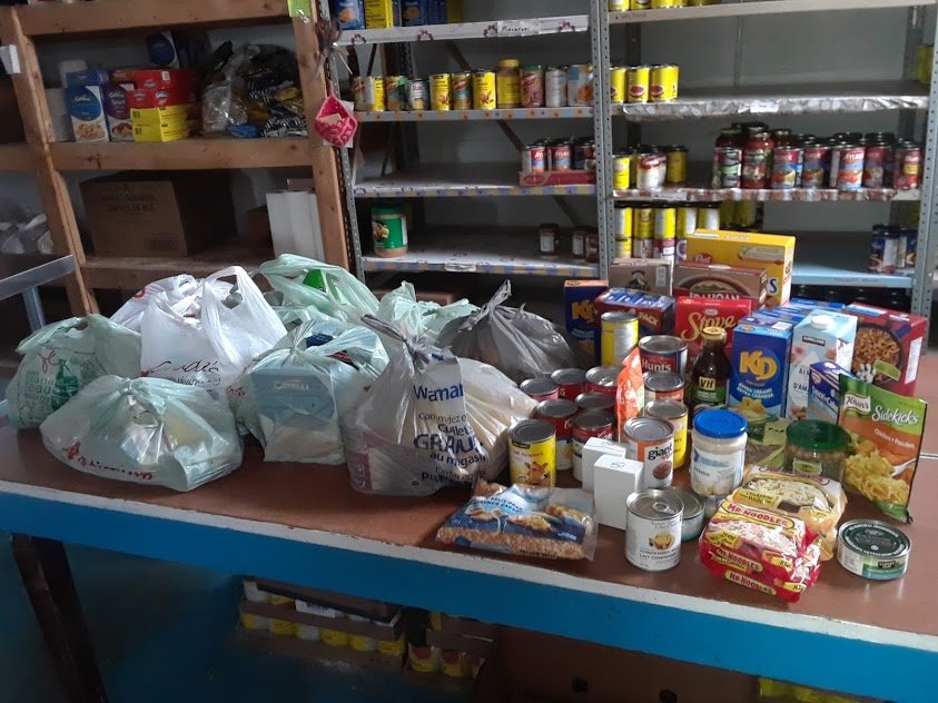 Image description: A table with full plastic grocery bags and canned and boxed food products is seen in the foreground. Shelves with more canned and boxed food are seen in the background. Some of the food products include Kraft Dinner, Mr. Noodles, tuna, and evaporated milk 