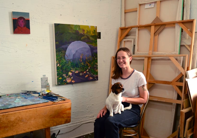 Lucy Koshan sits on a wood chair in her studio. There is a small white and brown dog in her lap. Canvas stretchers made of wood are behind her. A painting of a rock is mounted on the wall on the left.