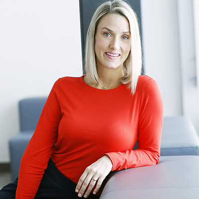 Maggie Brewer sits on a greyish blue chair. She is smiling. She is leaning on her left arm. She is white with blonde hair and blue eyes. She is wearing a red long-sleeved shirt. The room is brightly lit.