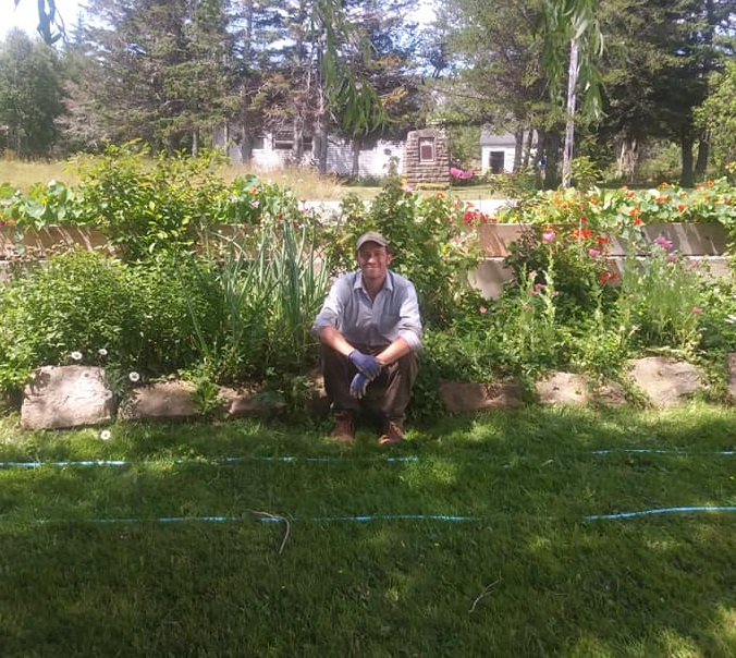 Image description: Michael Freeman sits in front of a garden. He is wearing gardening gloves and a hat, and is smiling. There are plants growing in wood and stone boxes. Some of the plants include poppies, nasturtiums, onions, and chives. There is a hose in front of Michael 