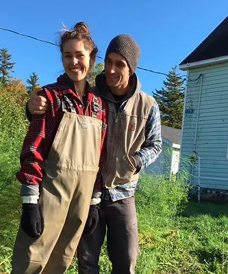 Sarah Smith and Jett Belliveau stand next to each other. Jett has his hand on Sarah's shoulder. Sarah is white with long brown hair in a bun. She is wearing a red plaid shirt with brown work overalls. Jett is white and wearing a toque, a brown vest, a plaid shirt, and work vest. They are outdoors. The grass is green and there are no clouds in the sky. It is daytime.