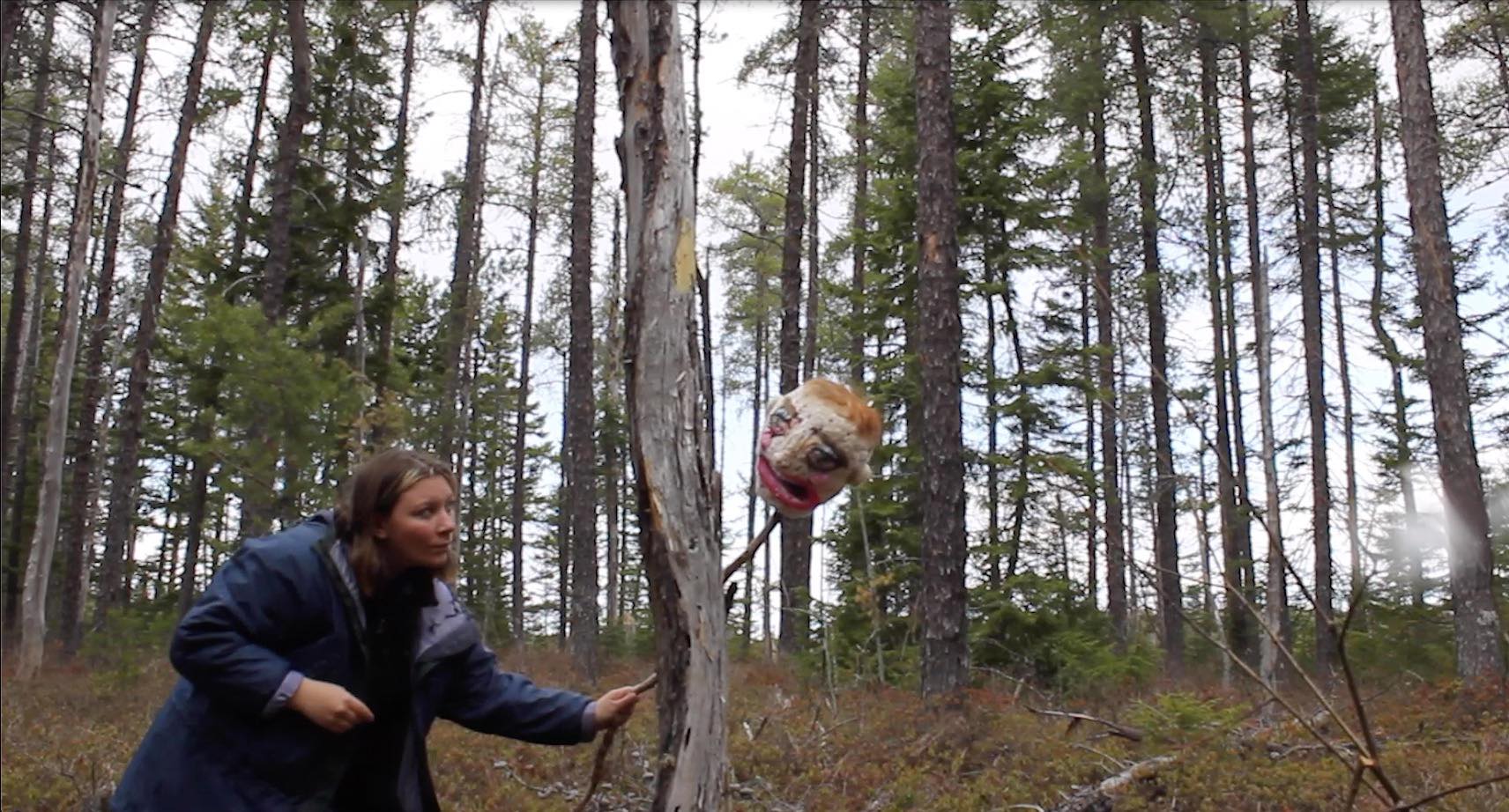 Emma Delaney holds a felted puppet head, larger than a human head, at the end of a stick. The stick goes behind a tree. Multiple pine trees are in the background. It is daytime.