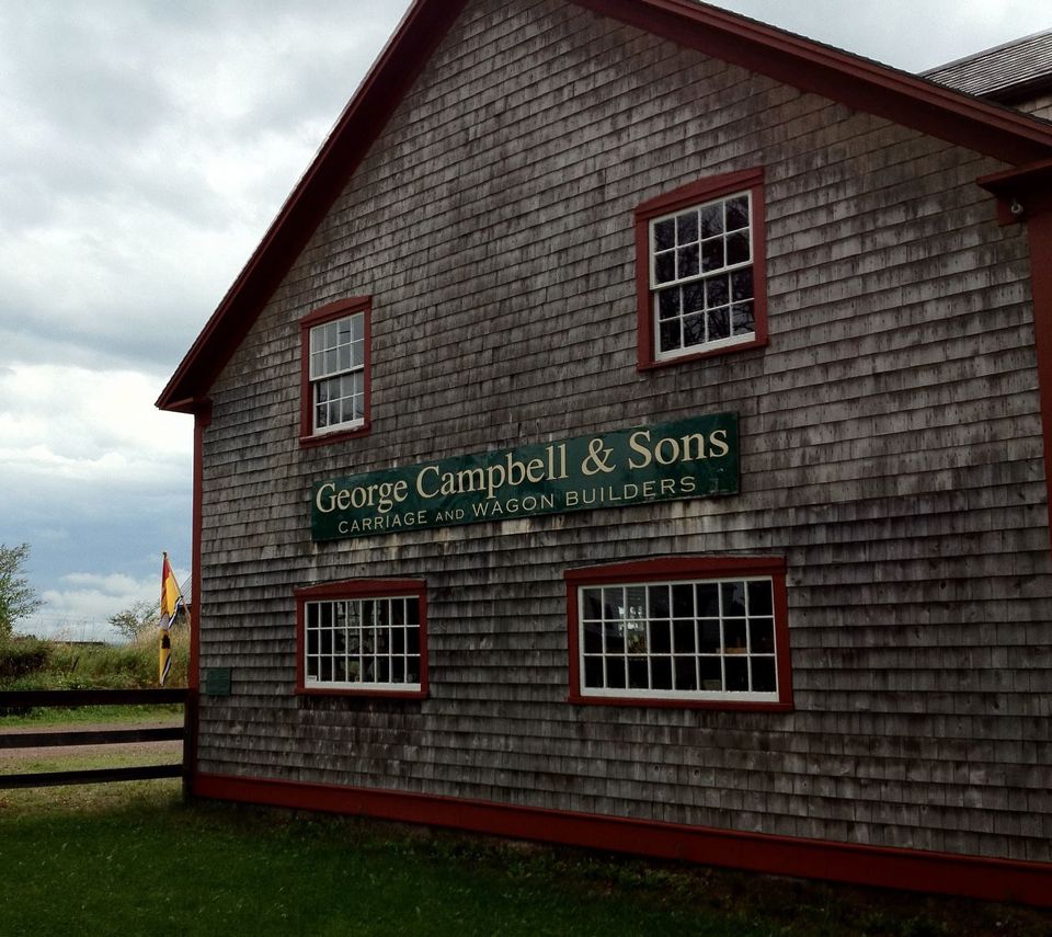 The side of the Campbell Carriage Factory Museum is shown. The building is shingled and the trim around the windows and frame is red. The window panes are painted white. The grass is green. It is daytime.