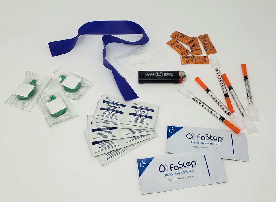 Safe injection supplies are displayed on a lightbox. There is a tourniquet, five needles, alcohol swabs, and a lighter. 