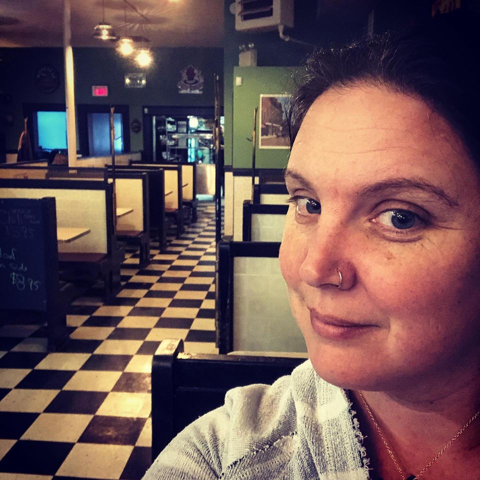 Ashley Legere stands in front of restaurant booths. The floor is black and white tiles. The photo is filtered. Ashley is white with blue eyes, brown hair, and a nose ring.