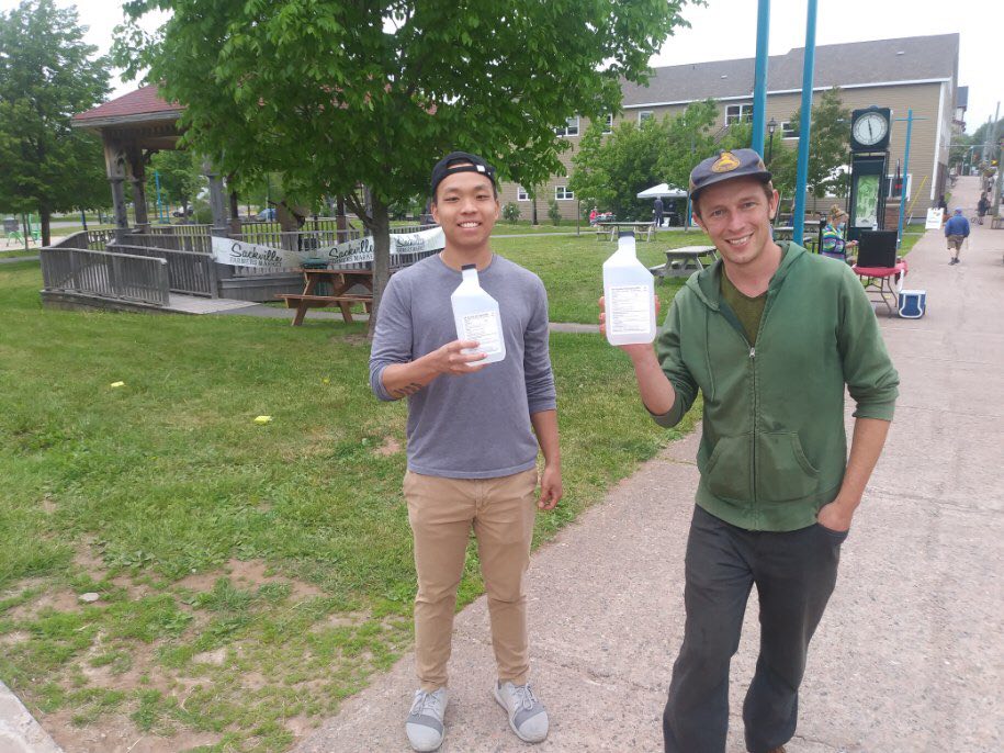 William Njo and Michael Freeman stand outside on the sidewalk. They are each holding a bottle of hand sanitizer. The farmers' market is ongoing in the background.