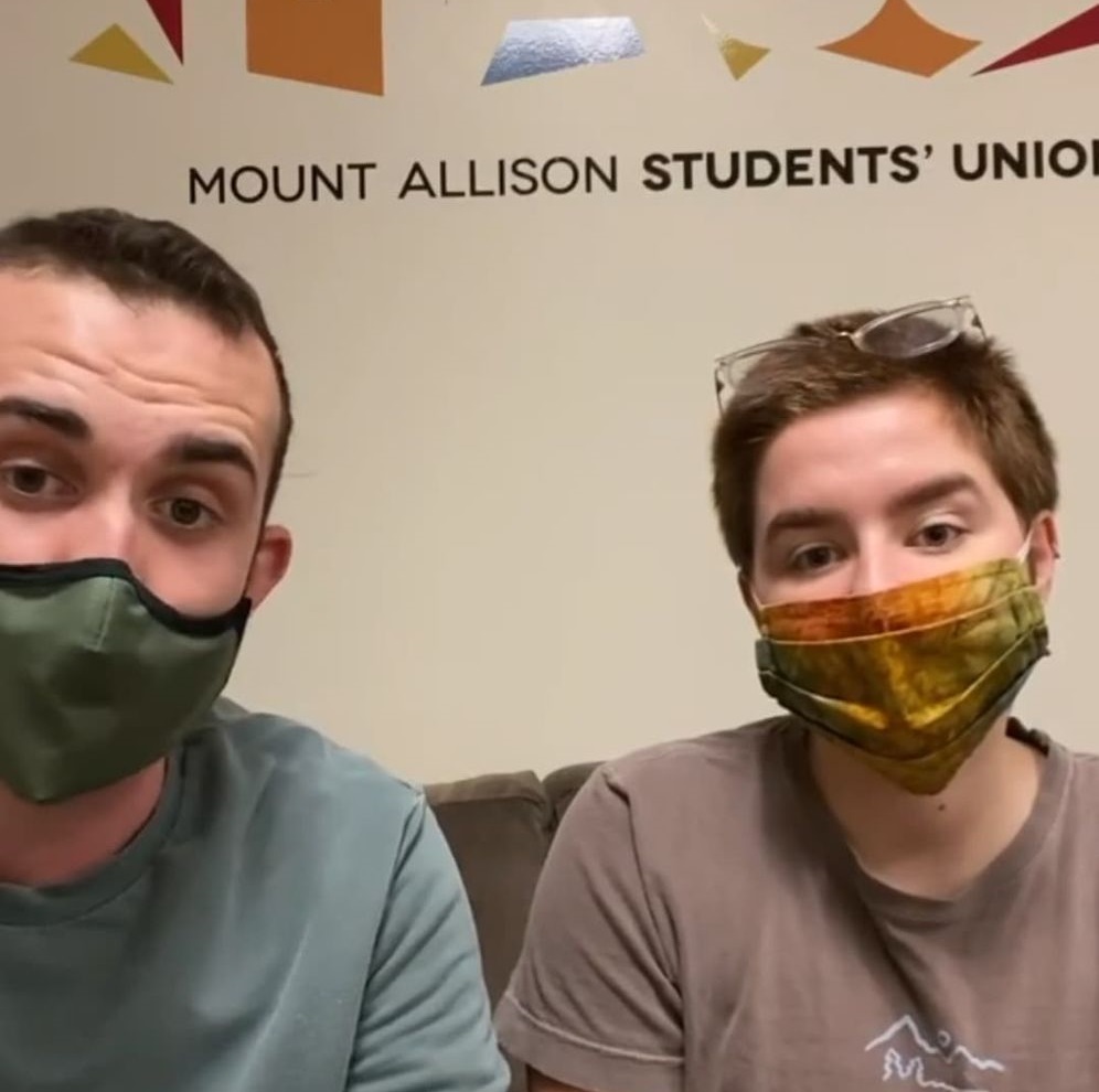 Jon Ferguson and Charlie Burke sit side by side on a couch with face masks on.