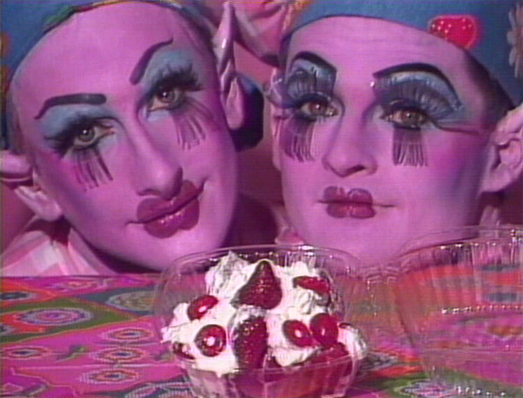 Two people in drag rest their chins on a table with whipped cream and strawberries in a container in front of them.