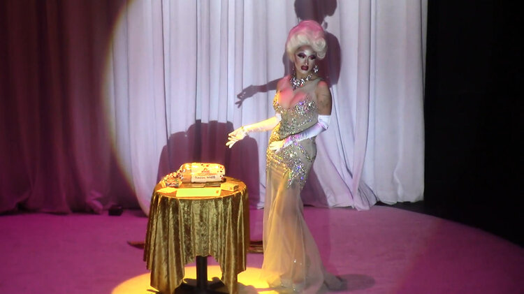 A person in drag wearing white gloves and a sequined dress gestures to a table with bread on it.