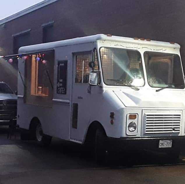 A white food truck is parked beside a brick building and is decorated by lights.