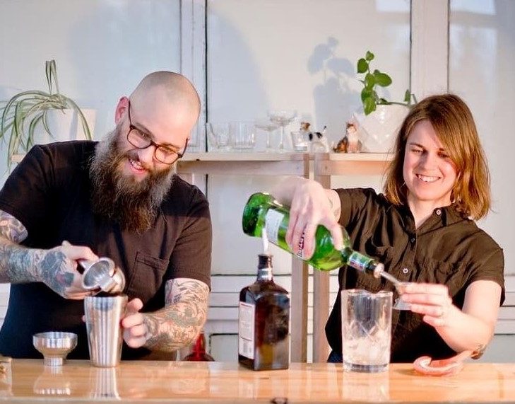 Glenn Barrington and Anne Herteis make drinks behind a bar, side by side.