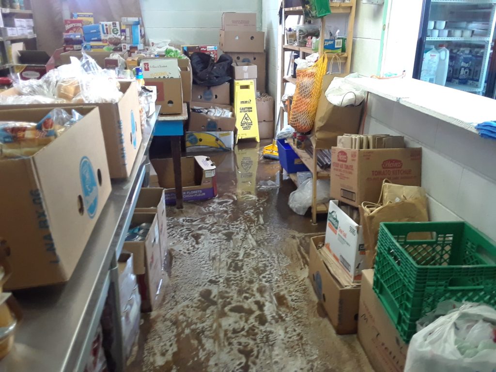 Cardboard boxes of food sit on stainless steel counters and shelves. The floor is covered in suds and water.