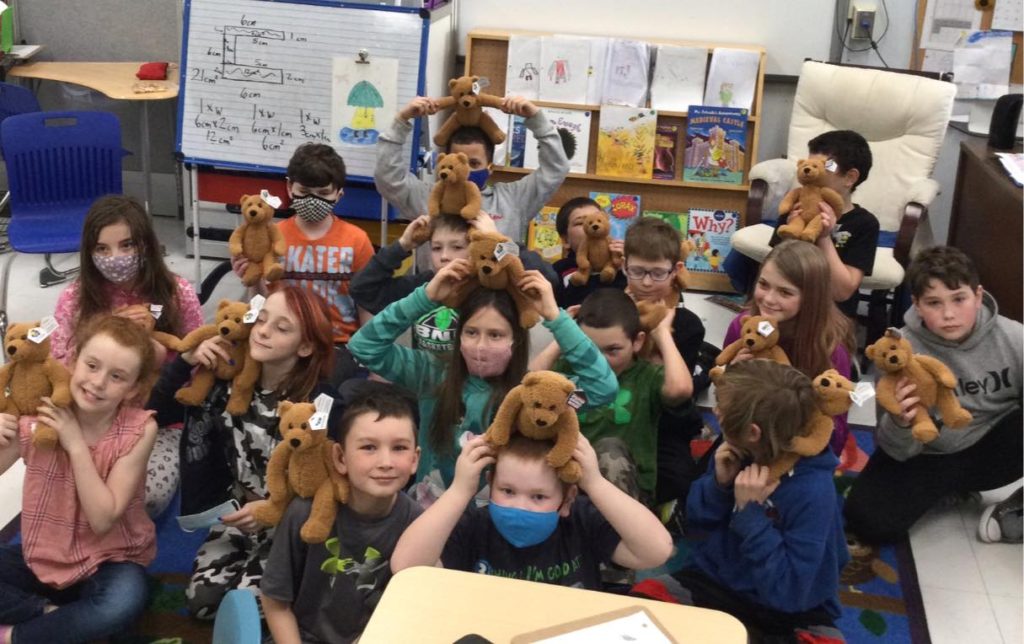 A classroom full of kids holding identical teddy bears and smiling.