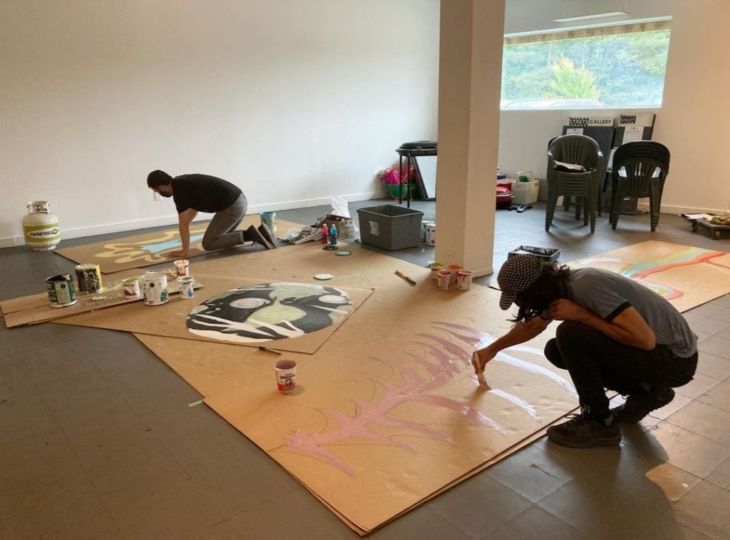 Two people crouch on the floor and paint a large brown sheet of paper.