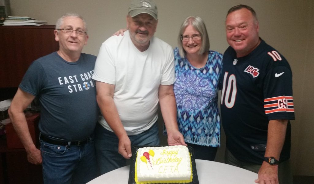 Four people stand around a table with a cake on it and smile.