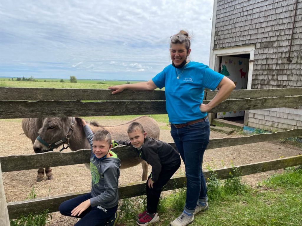 Laura Hunter leans on a fence with her two small grandchildren. There is a donkey behind the fence.