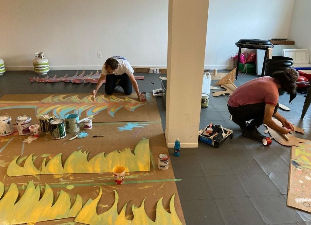 Two people kneel on the floor and paint yellow patterns onto brown paper.