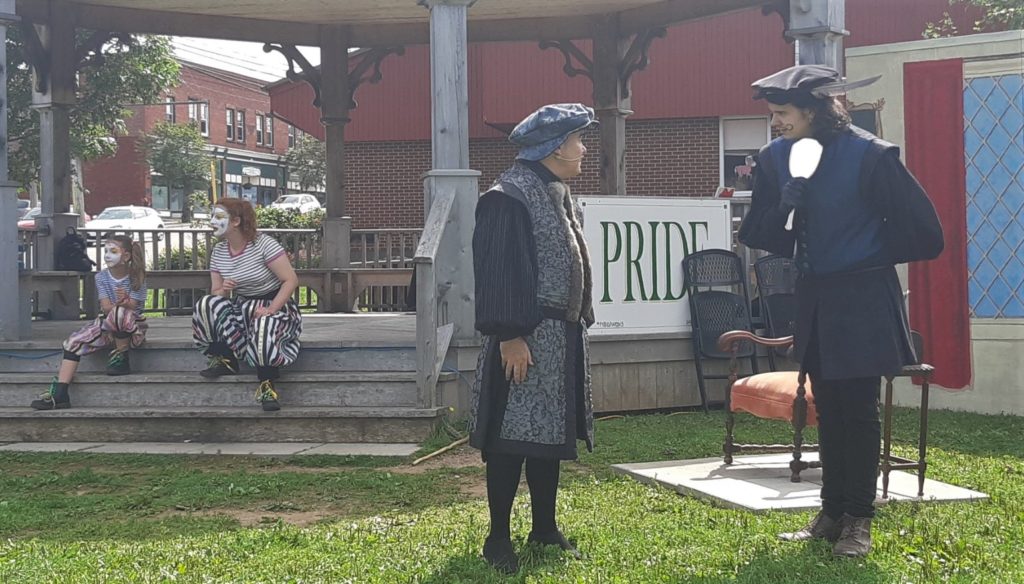 Four actors perform outdoors. Two are dressed as clowns and sit on the steps of a bandstand, while two are in medieval clothing and talk to each other on the grass.