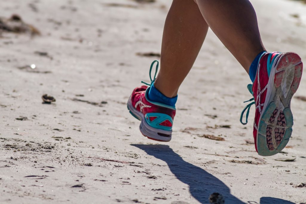 A person is seen from the knee down, running in sneakers on a beach.