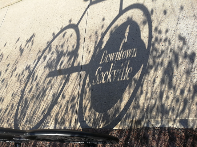 a shadow of a "Downtown Sackville" bike rack on a sidwalk