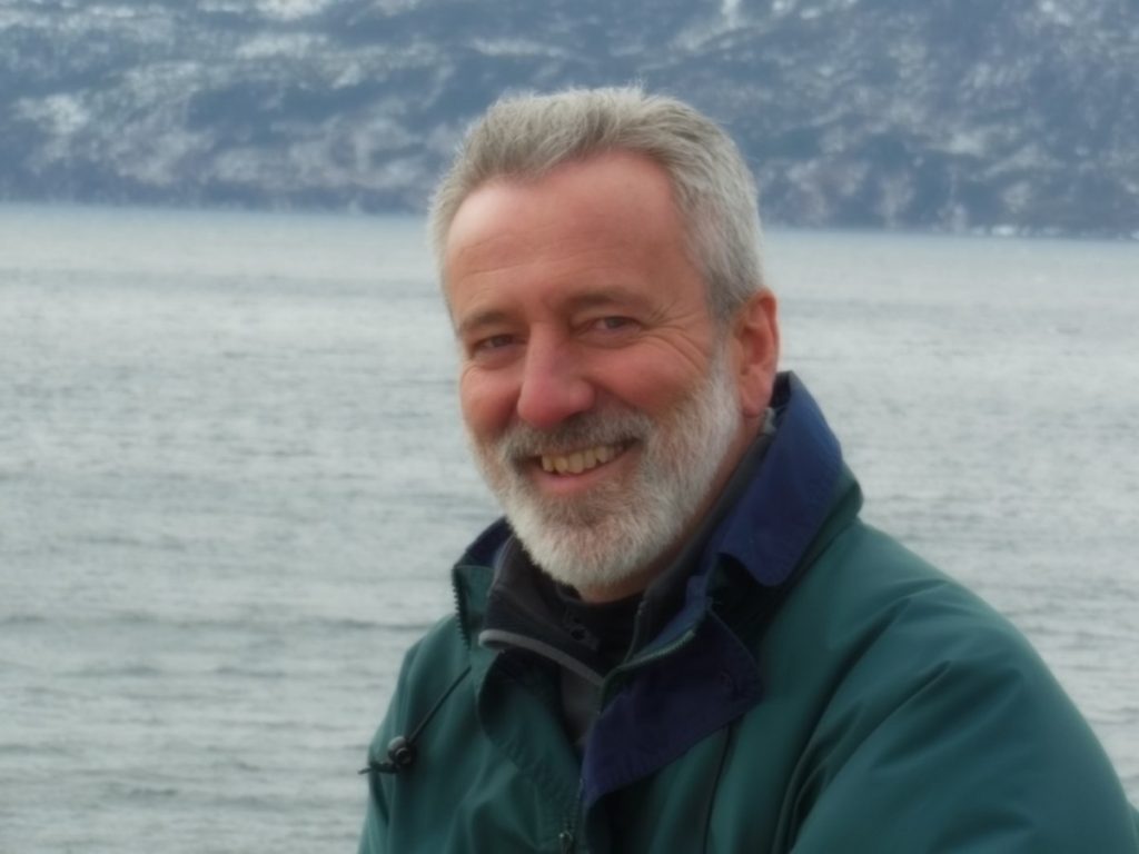 A bearded man is shown in a profile shot with a river or other body of water in the background. 