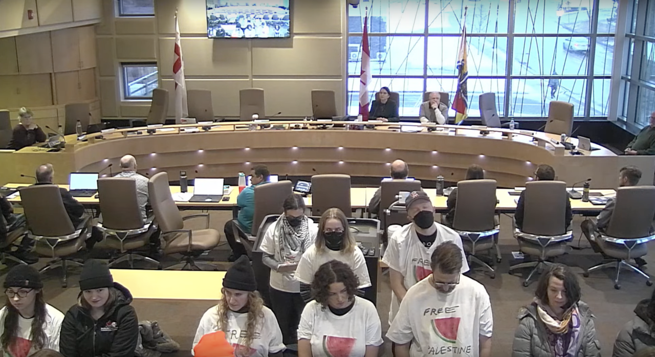 A council chambers with a number of people in white t-shirt facing the camera, and away from the council table, where many of the seats are empty.