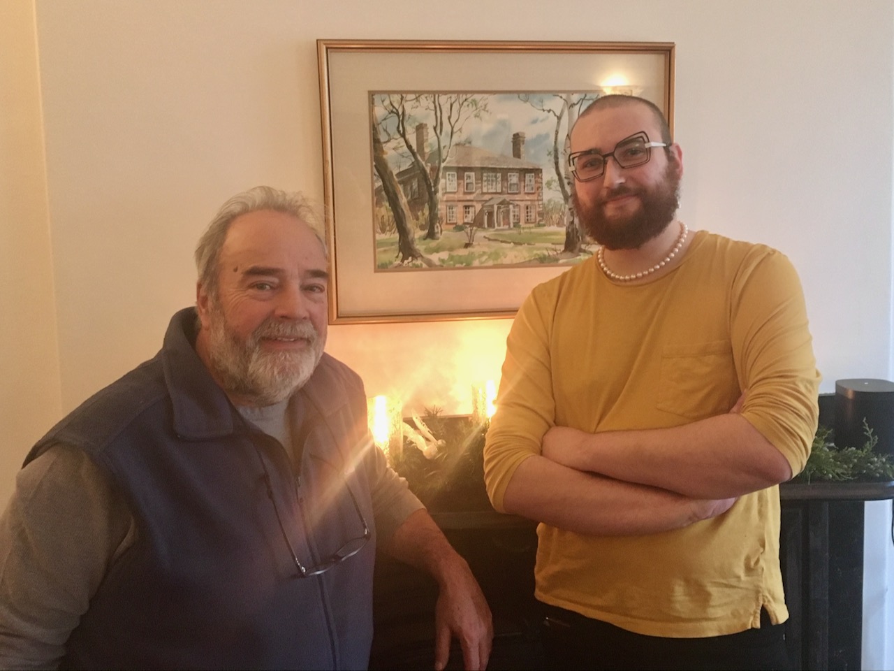 An older, shorter man and a taller younger man stand in front of a mantel piece, with a painting of a historic home hanging behind them.