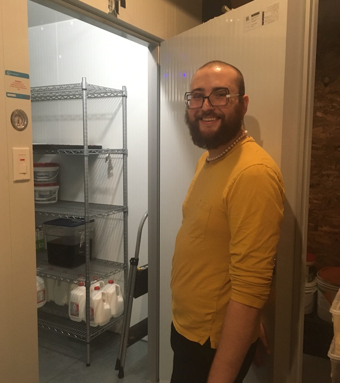 A man standing at an open door to a walk in fridge with shelving inside holding 4 litre jugs of milk. 